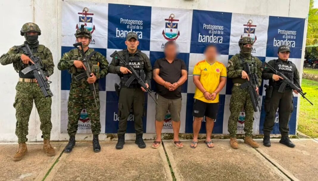 Efectivos armados custodian a dos detenidos con el rostro pixelado frente a un panel institucional
