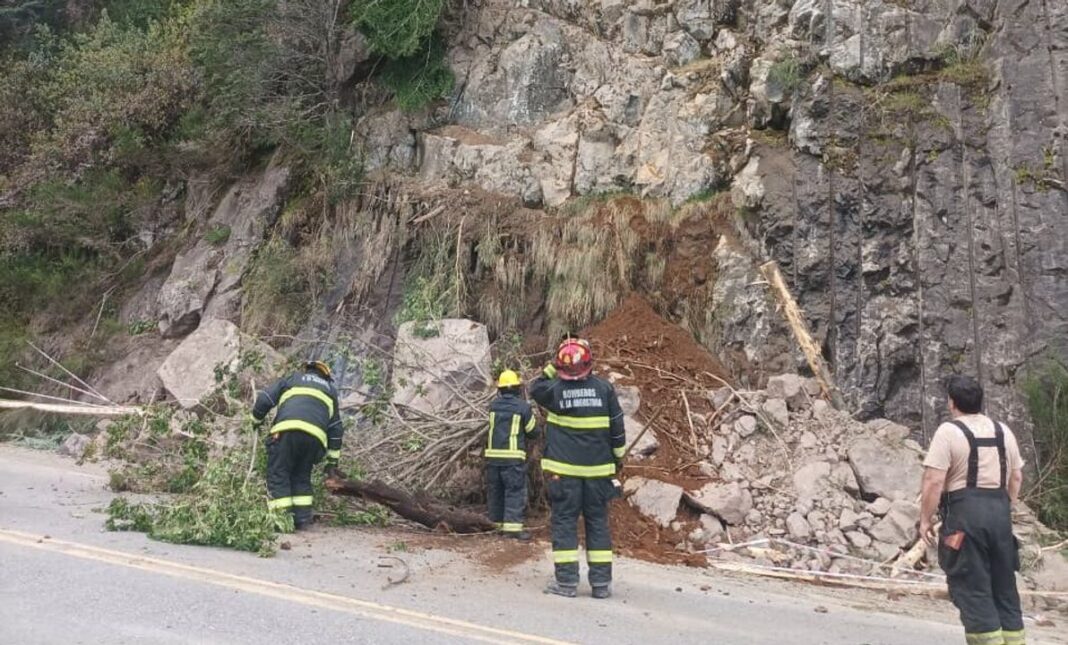 Bomberos trabajan junto a rocas, tierra y árboles caídos sobre una ruta al pie de una ladera rocosa
