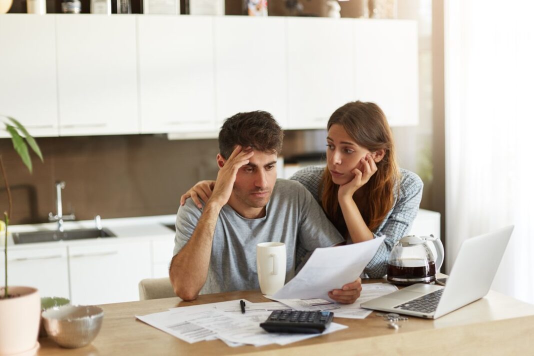 Una pareja preocupada revisa facturas y papeles en la mesa de la cocina, con una calculadora