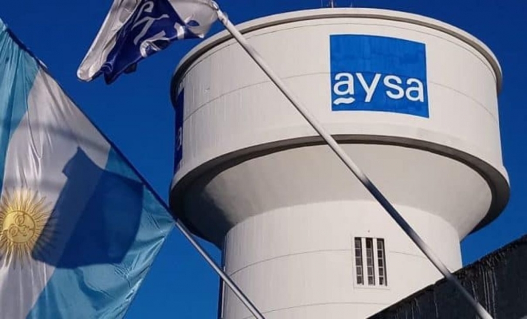 AySA se privatiza: Rowing y Roggio pujan por el agua del AMBA con tarifas en revisión Tanque de agua con el logo de AySA junto a una bandera argentina